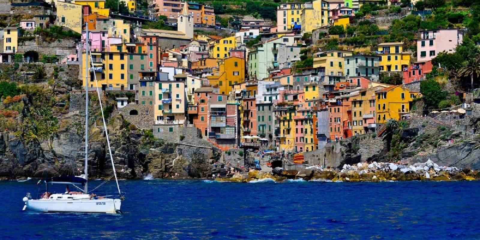 Pastel cliffside village overlooking a bright blue sea with a sailboat in the foreground, evoking Mediterranean travel
